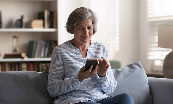 Eine Frau sitzt auf ihrem Sofa und hat ein Smartphone in der Hand. Im Hintergrund sieht man ein Bücherregal.