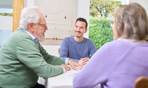 Zwei ältere Menschen – ein Mann und eine Frau – sitzen mit einem Pflegeberater am Tisch. Auf dem Tisch liegen Papiere.
