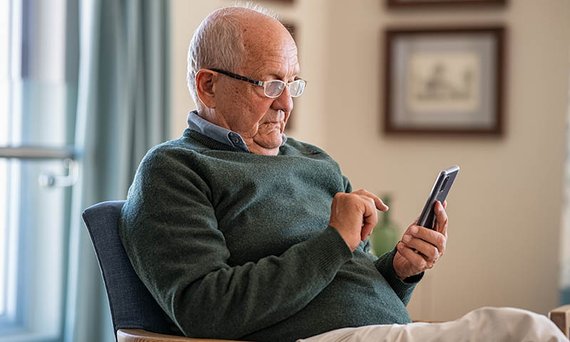 Ein älterer Mann mit Lesebrille sitzt in einem Sessel und hält ein Smartphone in der Hand.  