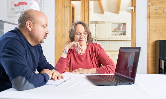 Eine Frau und ein Mann mit Notizblock sitzen an einem Tisch vor einem Laptop.