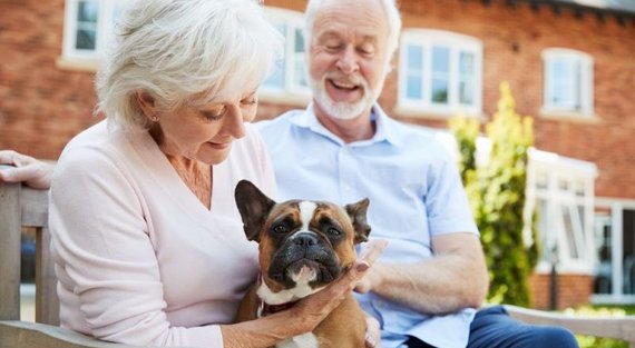 Senior und Seniorin sitzen mit Hund auf einer Bank vor einem mehrgeschossigen Wohnhaus. 