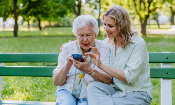Zwei Personen sitzen auf einer Parkbank. Die jüngere zeigt der älteren etwas auf dem Smartphone.
