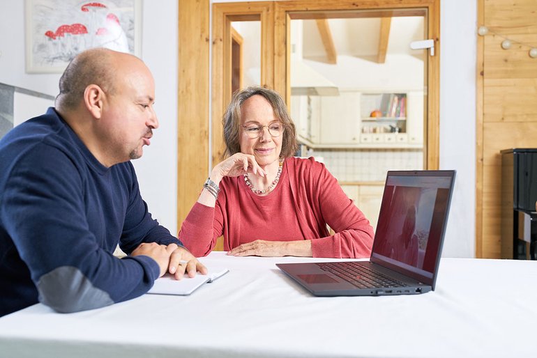 Eine Frau und ein Mann mit Notizblock sitzen an einem Tisch vor einem Laptop.