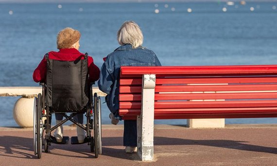 Zwei Personen schauen nebeneinander aufs Meer, die eine im Rollstuhl, die andere auf einer roten Bank sitzend.