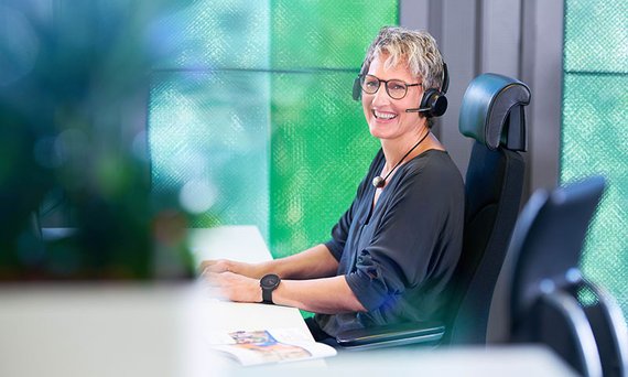 compass-Beraterin sitzt mit Headset an ihrem Schreibtisch und blickt freundlich in die Kamera.