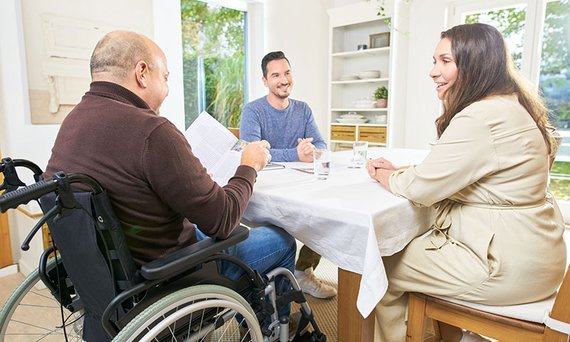 Ein Pflegeberater spricht an einem Tisch sitzend mit einem pflegebed&uuml;rftigen Mann im Rollstuhl und dessen Ehefrau.