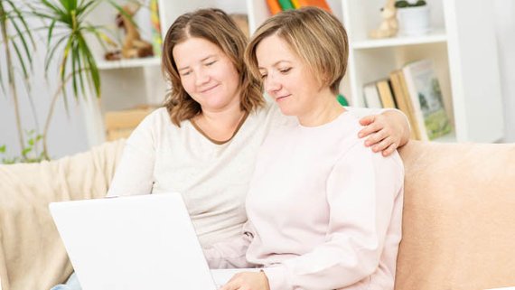 Zwei Frauen sitzen gemeinsam auf einer Couch vor einem Bücherregal. Sie haben einen Laptop auf dem Schoß, in den sie gemeinsam reinsehen. Eine Frau hat der anderen den Arm um die Schulter gelegt.