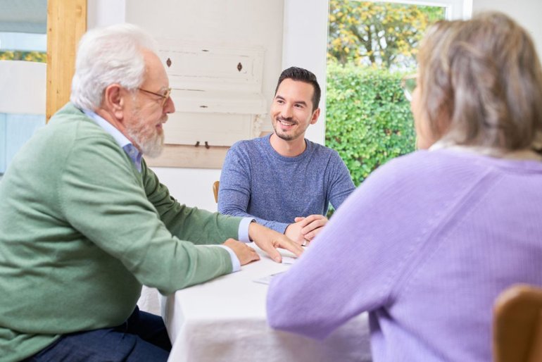Zwei ältere Menschen – ein Mann und eine Frau – sitzen mit einem Pflegeberater am Tisch. Auf dem Tisch liegen Papiere.