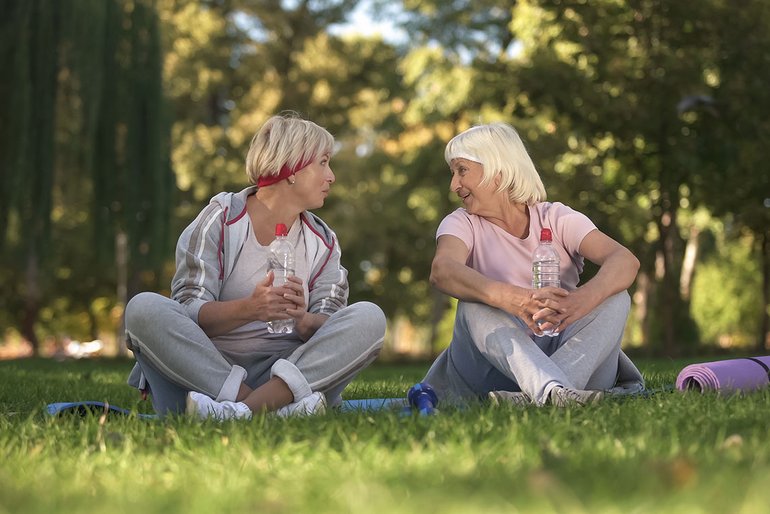 Zwei sportlich gekleidete Frauen sitzen nebeneinander im Gras. Sie sind einander zugewandt, sprechen miteinander und lächeln. Sie halten Trinkflaschen in den Händen. Im Hintergrund sind Bäume zu sehen.