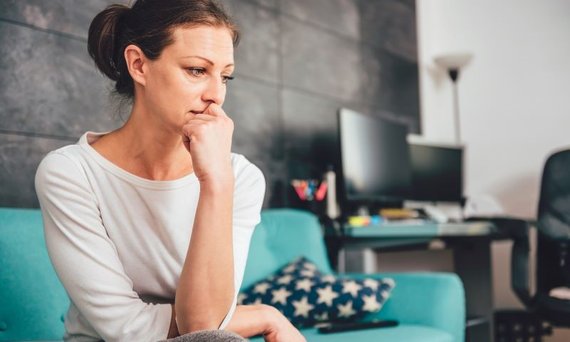 Frau mittleren Alters sitzt mit abgewendetem Blick auf einem Sofa. Im Hintergrund ist ein Schreibtisch mit zwei Bildschirmen zu sehen.