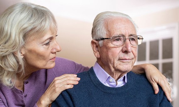 Eine ältere Frau hat von hinten die Hände auf die Schultern eines älteren Mannes gelegt.