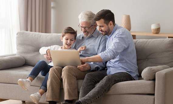 Drei Generationen einer Familie, Großvater, Sohn und Enkelkind, sitzen auf einem Sofa und schauen auf einen Laptop.