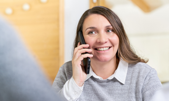 Eine Frau mittleren Alters hält ein Handy am Ohr und lächelt eine Person an, die ihr gegenübersitzt. Diese sieht man nur im Anschnitt.