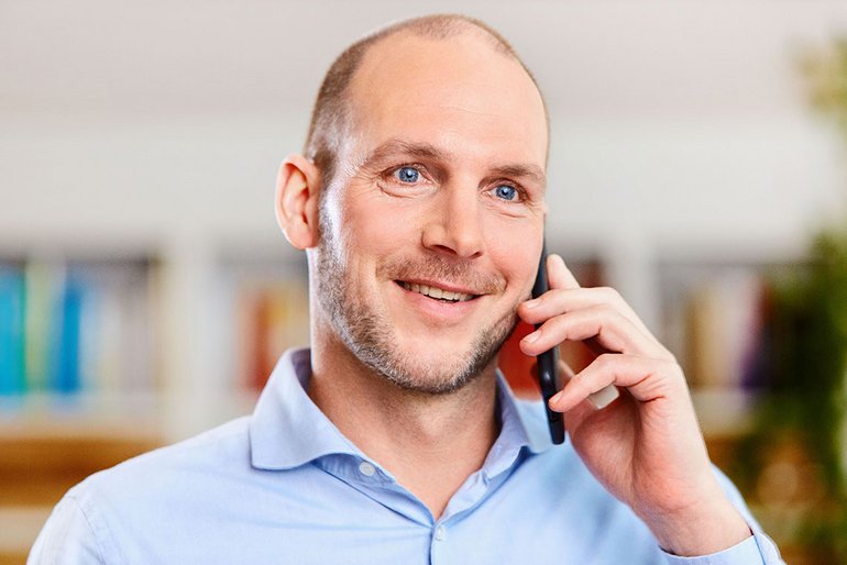 Ein Mann mit hellblauem Hemd steht im Fokus. Er schaut an der Kamera vorbei, der Mund ist geöffnet, halb lächelnd. Mit der linken Hand hält er ein Handy an sein Ohr.
