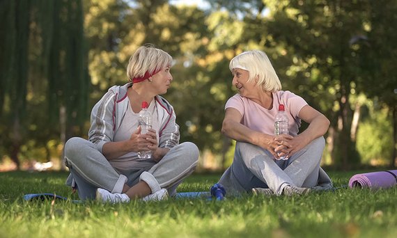 Zwei sportlich gekleidete Frauen sitzen nebeneinander im Gras. Sie sind einander zugewandt, sprechen miteinander und lächeln. Sie halten Trinkflaschen in den Händen. Im Hintergrund sind Bäume zu sehen.