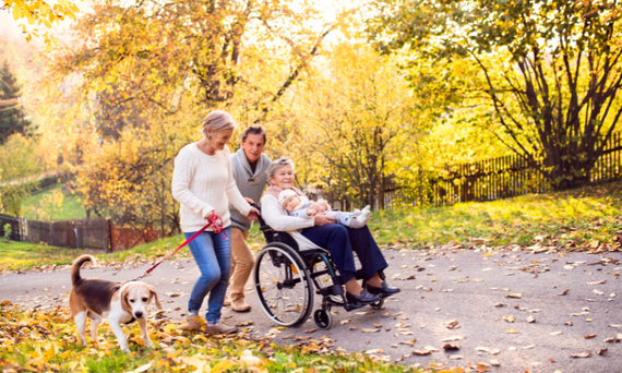 Eine Frau mit einem Hund an der Leine und ein Mann schieben eine Frau im Rollstuhl einen Weg entlang.