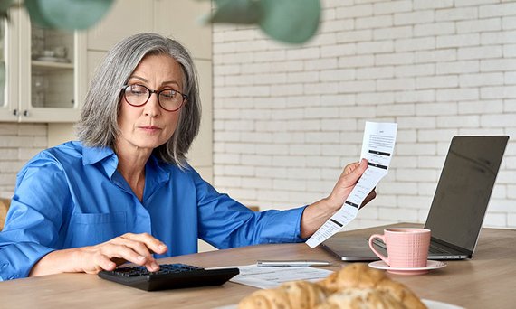 Eine Frau sitzt mit Dokumenten, Laptop und Taschenrechner am Frühstückstisch.