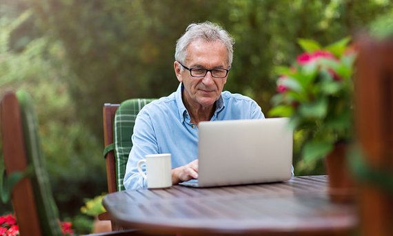 Ein Mann sitzt auf einem Holzstuhl mit Auflage am Gartentisch und macht eine Eingabe in einen Laptop. Neben ihm steht eine Tasse.